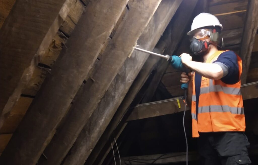 A person wearing an orange high-vis, hard hat, and face mask drilling into a wooden roof beam