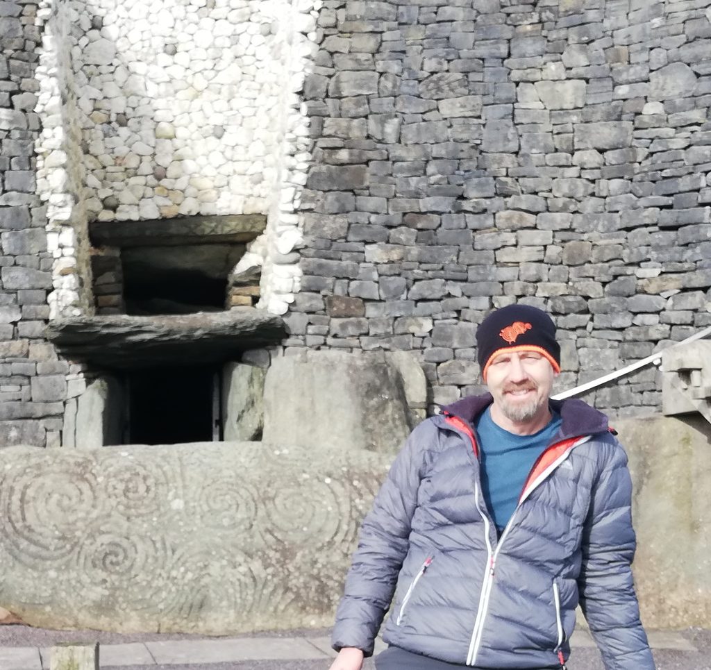 Photo of Gavin standing next to pictish  carved stone and in front of a stone wall